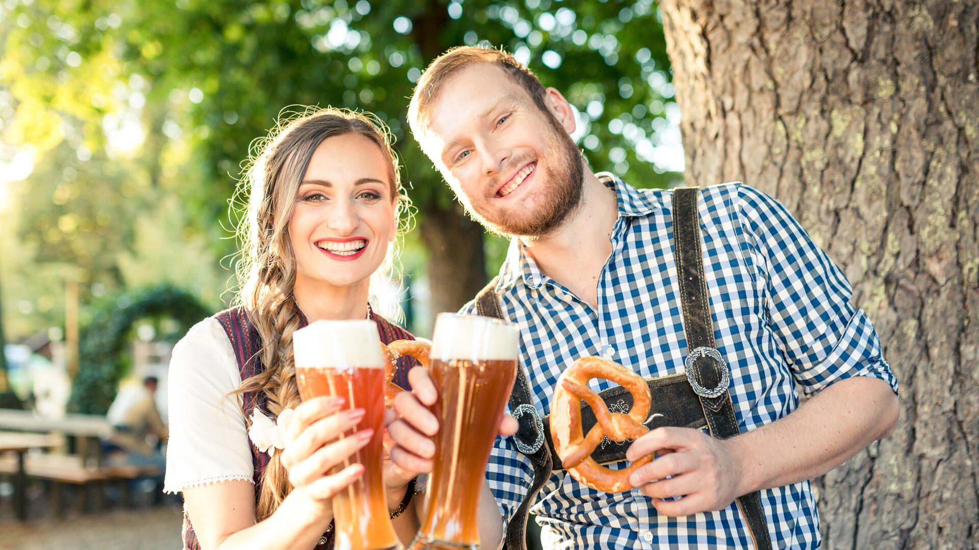 Paar in Tracht sitzt im Biergarten und prostet mit einem Weizenbier in die Kamera