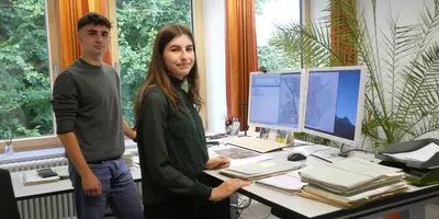 Johannes Fischer und Lena Vogl stehen jetzt als Katastertechniker vor ihrem Schreibtisch und lächeln in die Kamera. Foto von ADBV Straubing