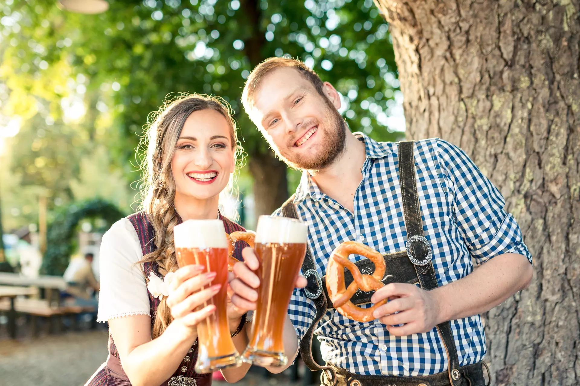 Stichwort „saisonal bedingt“ Paar in Tracht sitzt im Biergarten und prostet mit einem Weizenbier in die Kamera